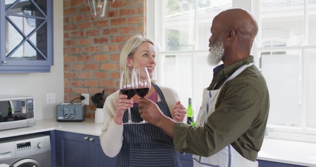 Mixed race senior couple wearing aprons toasting wine glasses while cooking in the kitchen at home. retirement senior couple lifestyle living concept