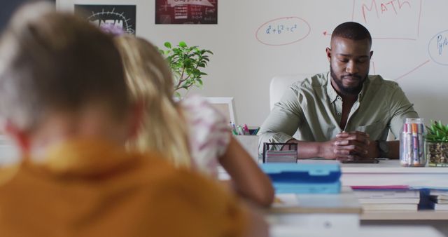 Image of happy african american male teacher sitting at desk during math lesson. primary school education and teaching profession.