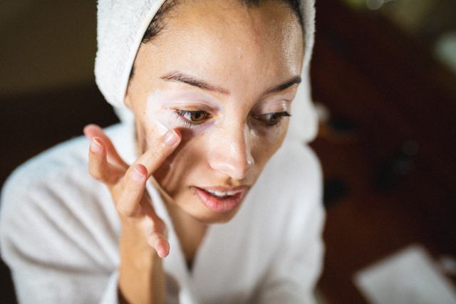 Close-up of biracial young woman in bathrobe applying cream on face during spa. unaltered, vitiligo, spa, skin care, body care, bathroom, lifestyle and wellness.