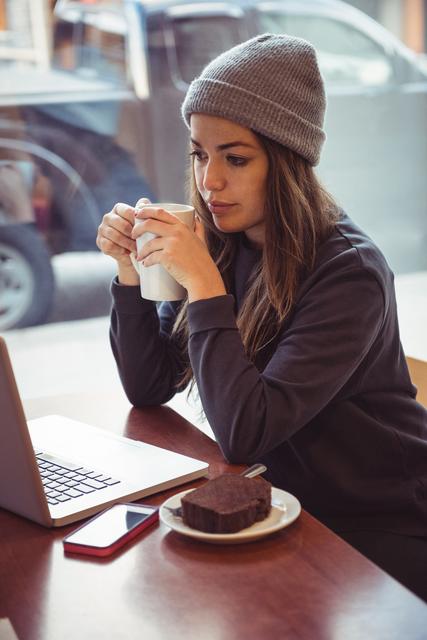 Woman in winter clothing holding coffee cup and looking at laptop in restaurant