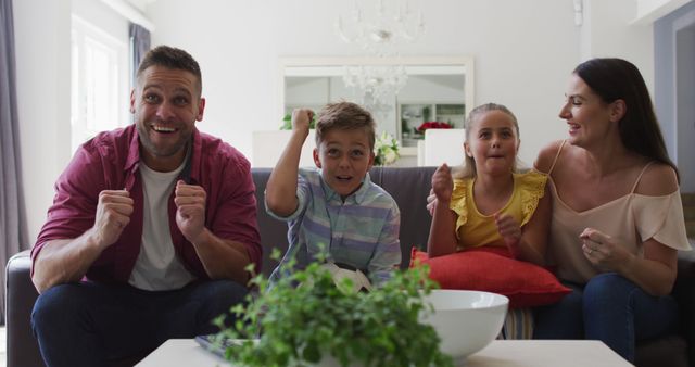 Excited caucasian parents, son and daughter on couch watching tv and cheering, son holding football. happy family, at home together.