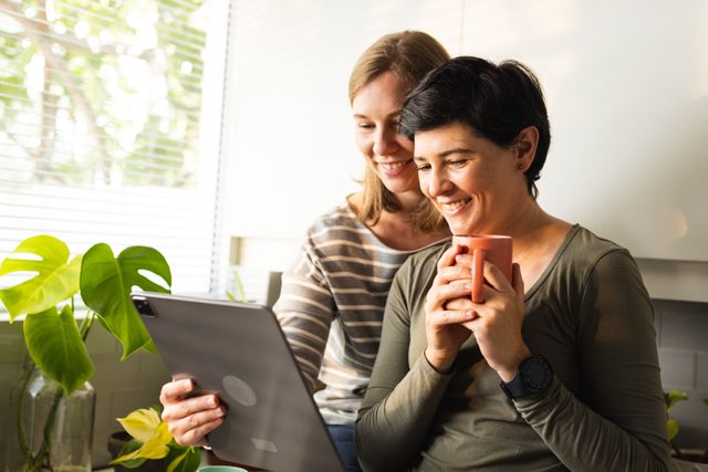 Smiling caucasian lesbian couple watching video over digital pc while having coffee in kitchen - Download Free Stock Photos Pikwizard.com