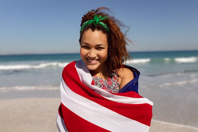 Portrait of beautiful young Mixed-race woman wrapped in american flag looking at camera on beach in the sunshine