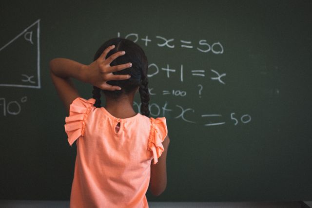 Rear view of mixed race schoolgirl standing at maths chalkboard in classroom holding head - Download Free Stock Photos Pikwizard.com