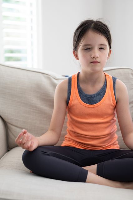 Little caucasian girl sitting on the couch with legs crossed meditating at home. she has her eyes closed.