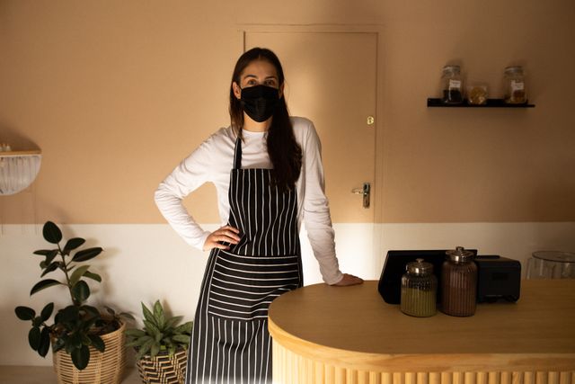 Portrait of caucasian female business owner wearing face mask and apron leaning on counter at cafe. independent cafe business owner during coronavirus covid 19 pandemic.